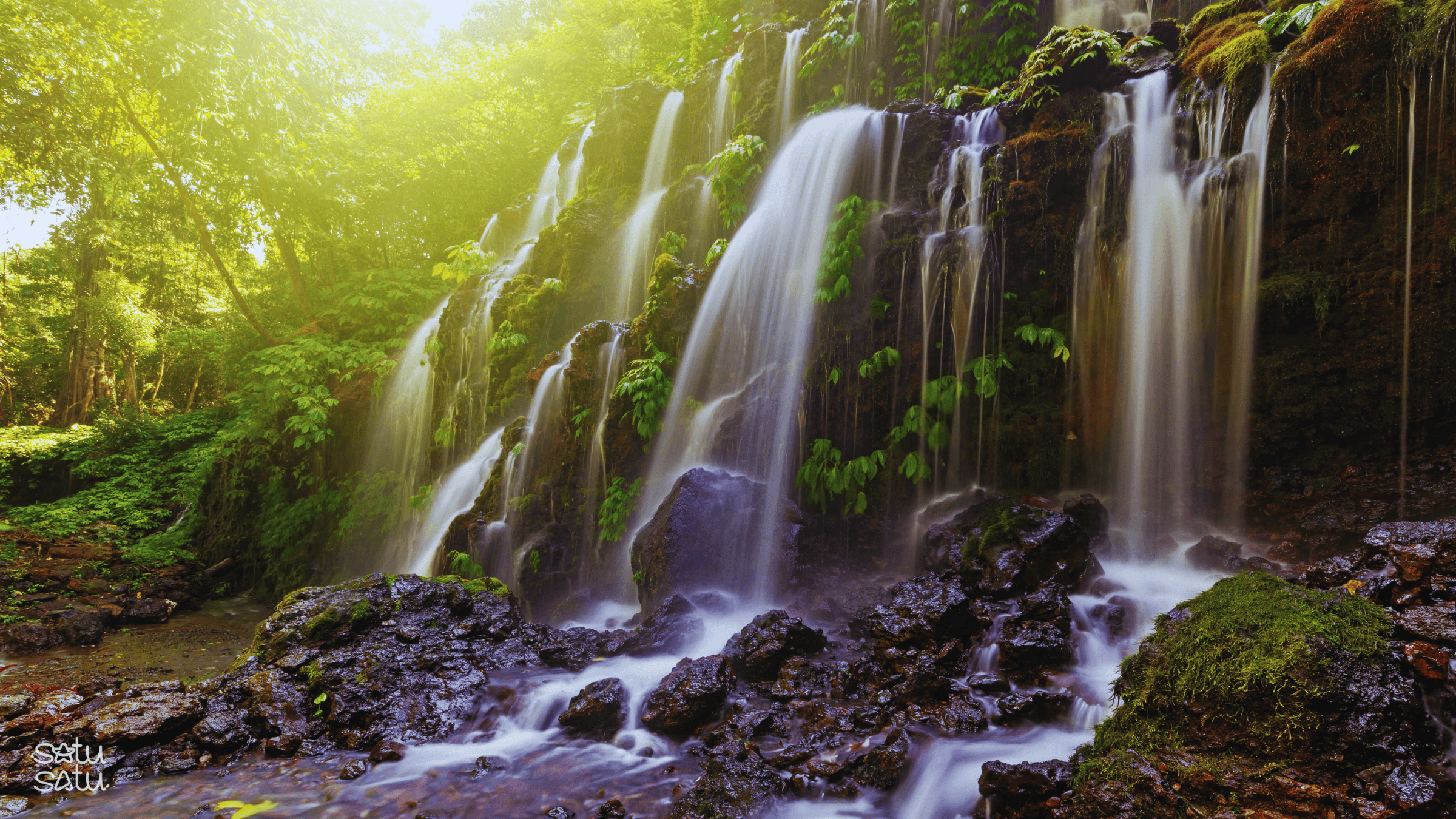 Banyuwana Waterfall in Bali with cascading streams surrounded by lush tropical forest and moss-covered rocks.