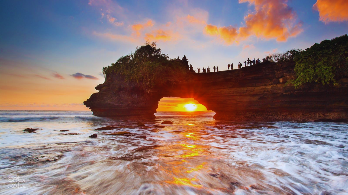 Sunset view at Pura Batu Bolong Bali with ocean waves, rock arch temple, and silhouettes of visitors watching the sunset in Bali, Indonesia.