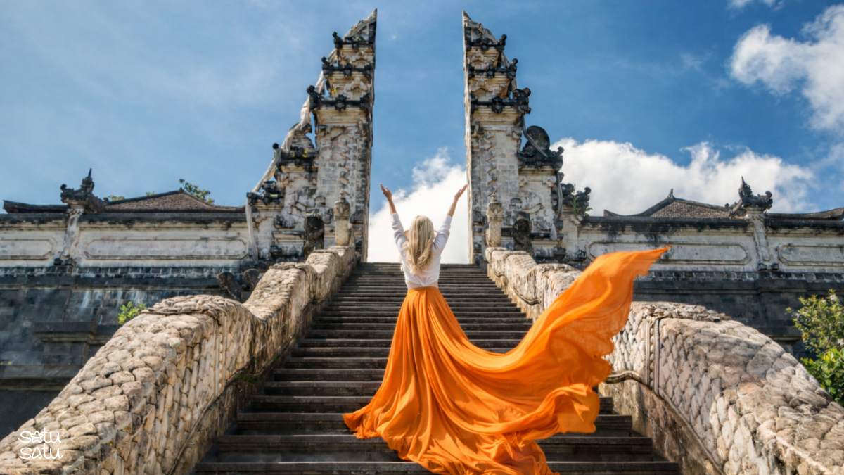 Woman standing on the stairs at Pura Lempuyang Bali with flowing orange dress, facing the iconic Gates of Heaven under blue sky.