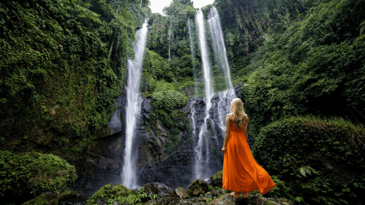 Traveler in an orange dress admiring the majestic Sekumpul Waterfall surrounded by lush tropical jungle in North Bali.