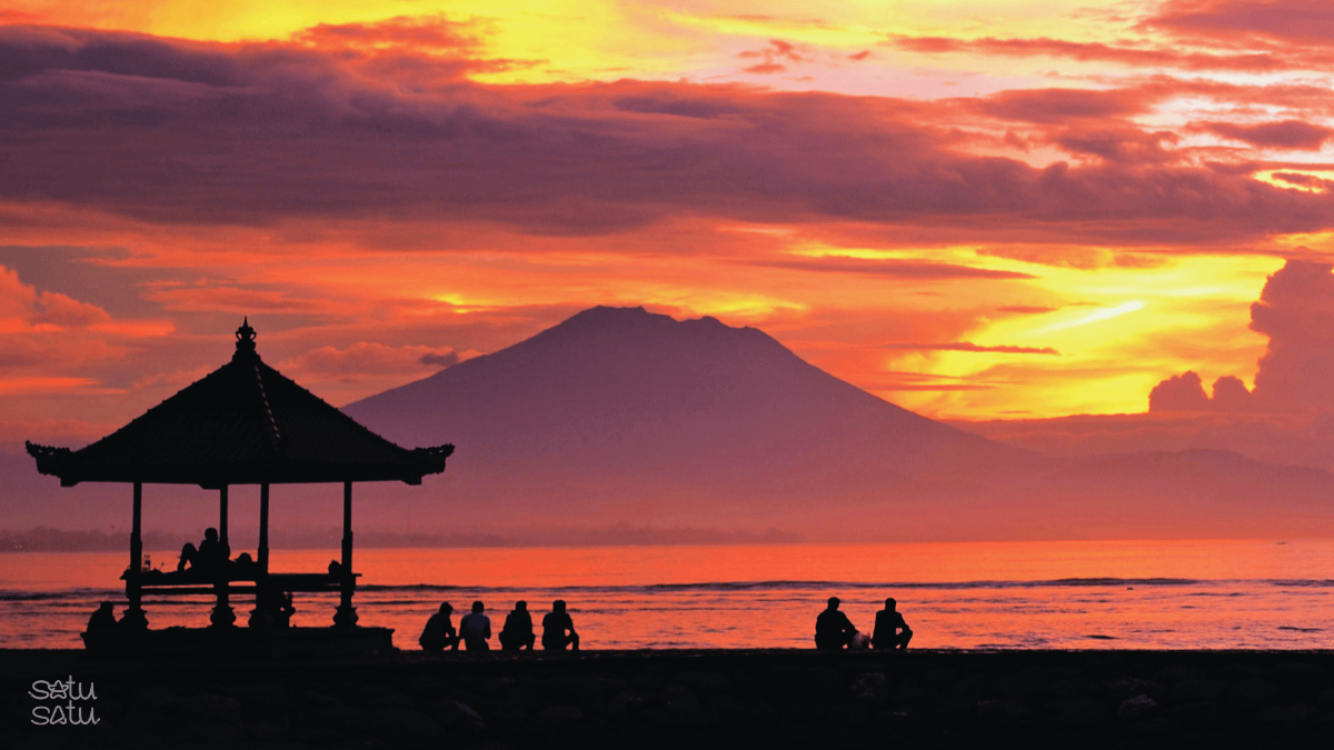 Sunrise at Sanur Beach Bali with Mount Agung silhouette, traditional gazebo, and people enjoying morning atmosphere by the sea.