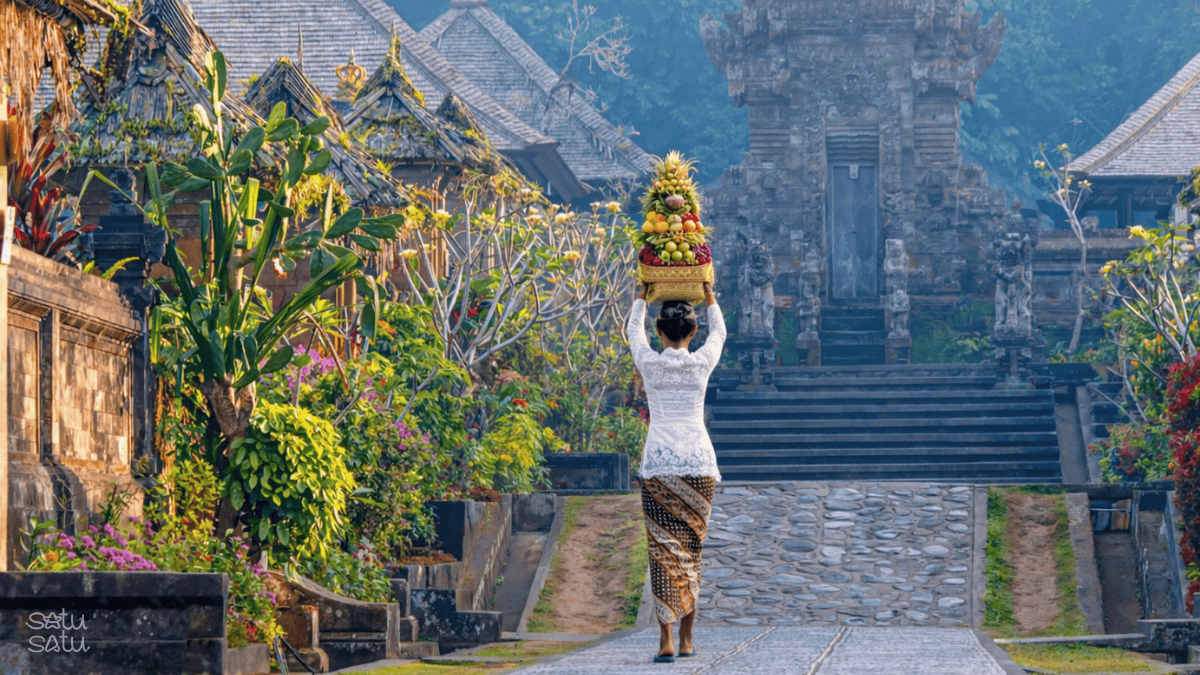 Balinese woman carrying traditional offerings in Tenganan Village, East Bali, surrounded by traditional houses and temple architecture.