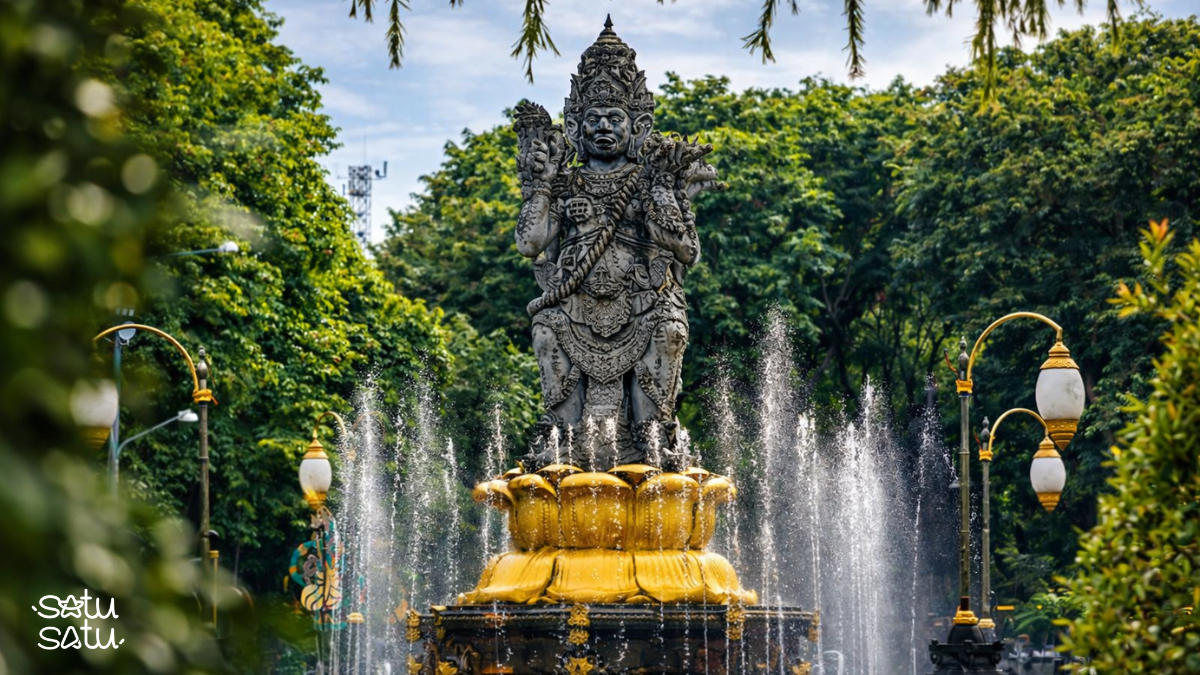 Caturmuka statue in Denpasar, Bali, standing on a fountain base surrounded by lush greenery and traditional decorative elements.