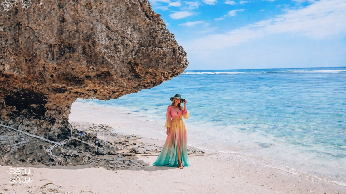 Female traveler standing on the shoreline of Thomas Beach in Bali near a large rock formation with clear blue ocean